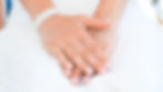 Bride's hands with a wedding ring, resting on a lace dress. She wears a pearl bracelet. The setting suggests a calm and joyful mood.
