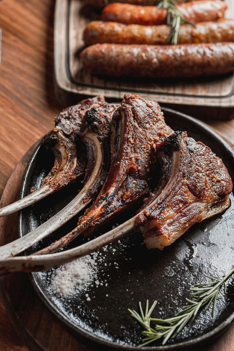 Grilled lamb chops on a dark plate with rosemary and salt. Background shows sausages on a wooden board. Warm, rustic setting.