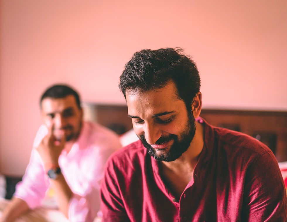 Two men inside a room, one in focus wearing a red shirt and smiling, the other in the background blurred. Warm lighting creates a cozy mood.