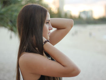 Woman with long brown hair stands on a beach, arms on head. She's wearing a black top and bracelet. Background shows blurred trees and buildings.
