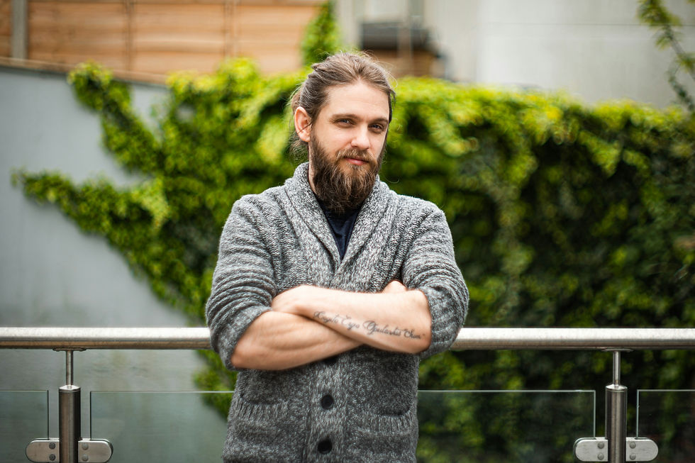 Bearded man in a gray cardigan stands with arms crossed on a balcony, in front of a blurred green plant wall.