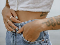 Person in white top and blue jeans, hands adjusting waistband. Visible tattoos and rings. Soft, blurred background. Casual mood.