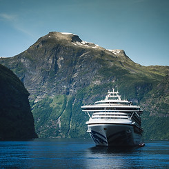 A majestic cruise ship anchored in a serene fjord with towering green mountains and a clear blue sky in the background.