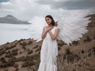 A woman in a white dress with angel wings stands on a grassy hill, hands clasped, under a cloudy sky, with a serene lake view.