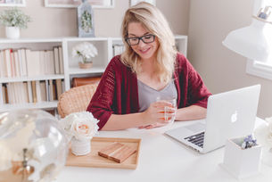 Woman smiling while holding a glass at a white desk with a laptop and flowers. Shelves and a window in the bright background.