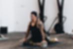 Man in a gym setting doing yoga, seated with yoga blocks and tattoos visible. Calm expression, wearing a black tank top and cap.