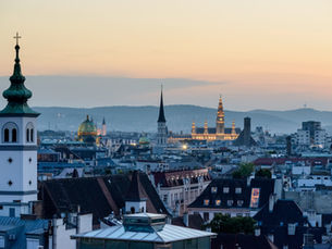 Cityscape at dusk with illuminated historic buildings, spires, and a green-domed structure. Mountains and a softly colored sky in the background.