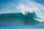 Surfer in a red shirt carves a huge blue wave, kicking up white spray under a clear sky.