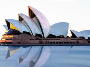 Sydney Opera House at sunset reflected in calm water, its white sail-like shells glowing against a clear sky.