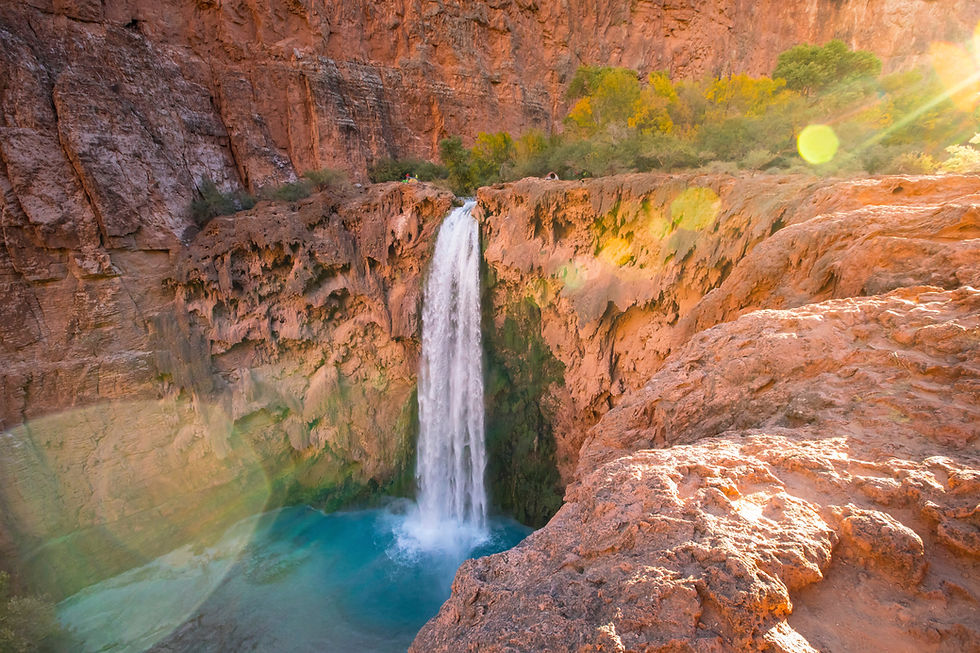 A waterfall cascades into a turquoise pool surrounded by red rock cliffs and trees, with sunlight creating lens flares in a tranquil setting.
