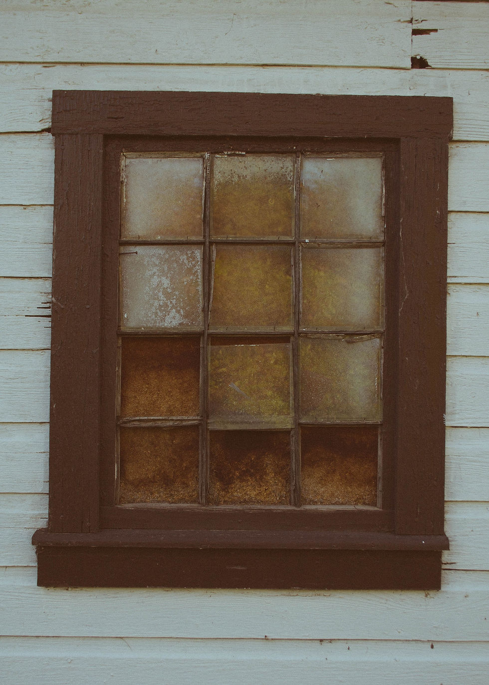 Weathered brown-framed window on peeling light blue wooden wall. Glass panes are aged and discolored, creating a rustic mood.