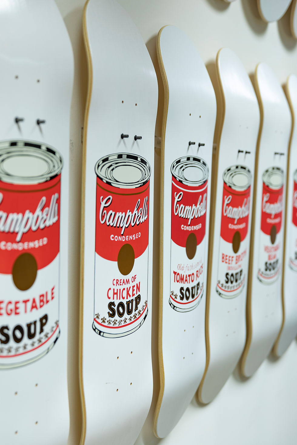 Skateboards with Campbell's soup can designs are lined up against a white wall. Bold red, white, and black colors dominate the scene.