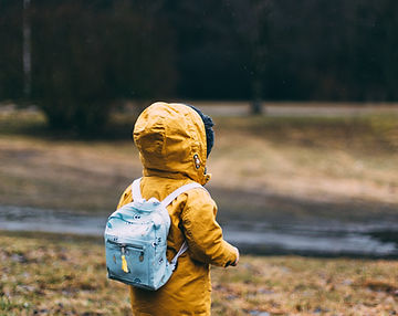 Child in a yellow coat with a backpack standing alone in a quiet park.