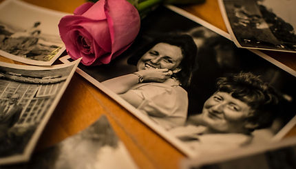 Pink rose resting on a table beside old black-and-white photographs of smiling women, symbolizing love, memory, and remembrance. Image by Laura Louise Grimsley