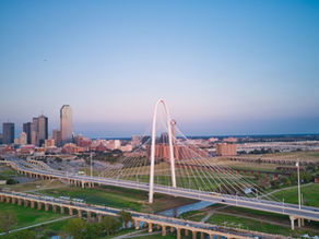 Aerial view of Dallas city skyline at sunset, featuring a prominent arched bridge over a green landscape. The sky is clear and blue.