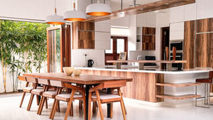 Modern open kitchen and dining area with wood accents, hanging pendant lights, long table and chairs, bright white floor, bamboo outside.