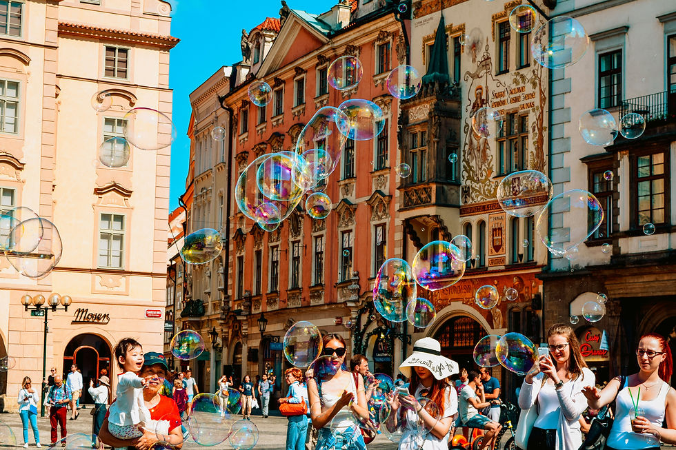 People in a sunny old city square blow giant soap bubbles, with ornate pastel buildings and a Moser storefront behind them.