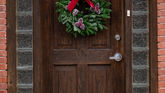 A wooden door with a festive wreath featuring a red bow and pinecones. Brick walls and glass panels frame the door, creating a cozy vibe.