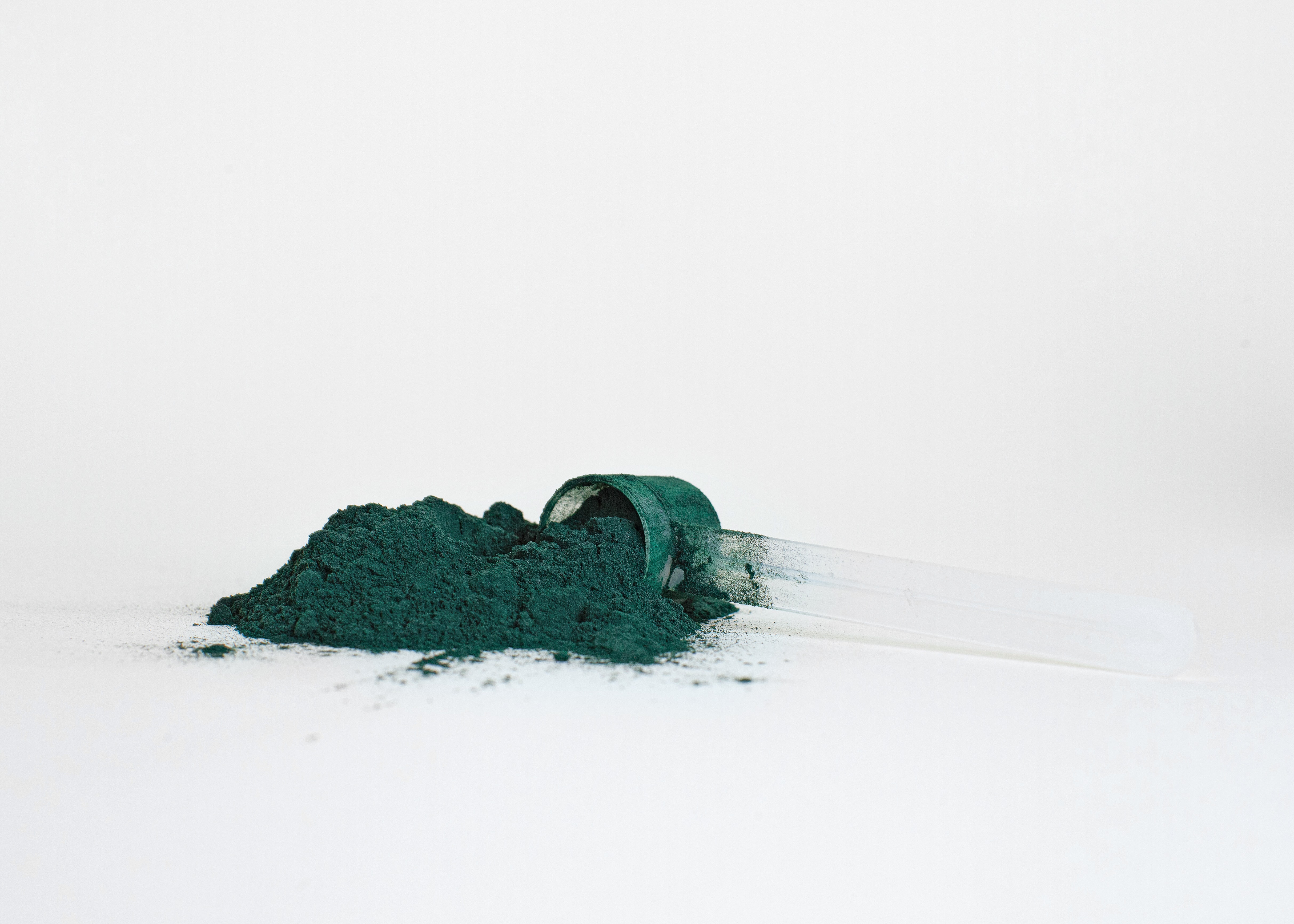 Pile of dark green spirulina powder spills from clear scoop on a white background.