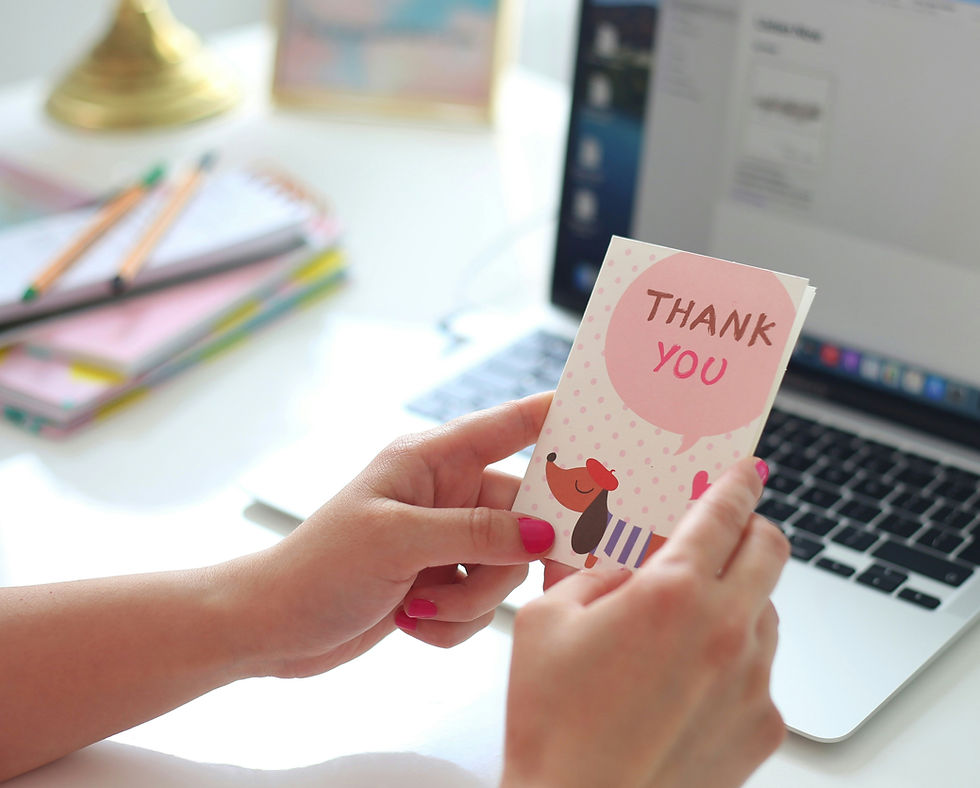 Hands holding a "Thank You" card with a dog illustration. Background shows a laptop and stationery on a white desk. Bright and cheerful mood.