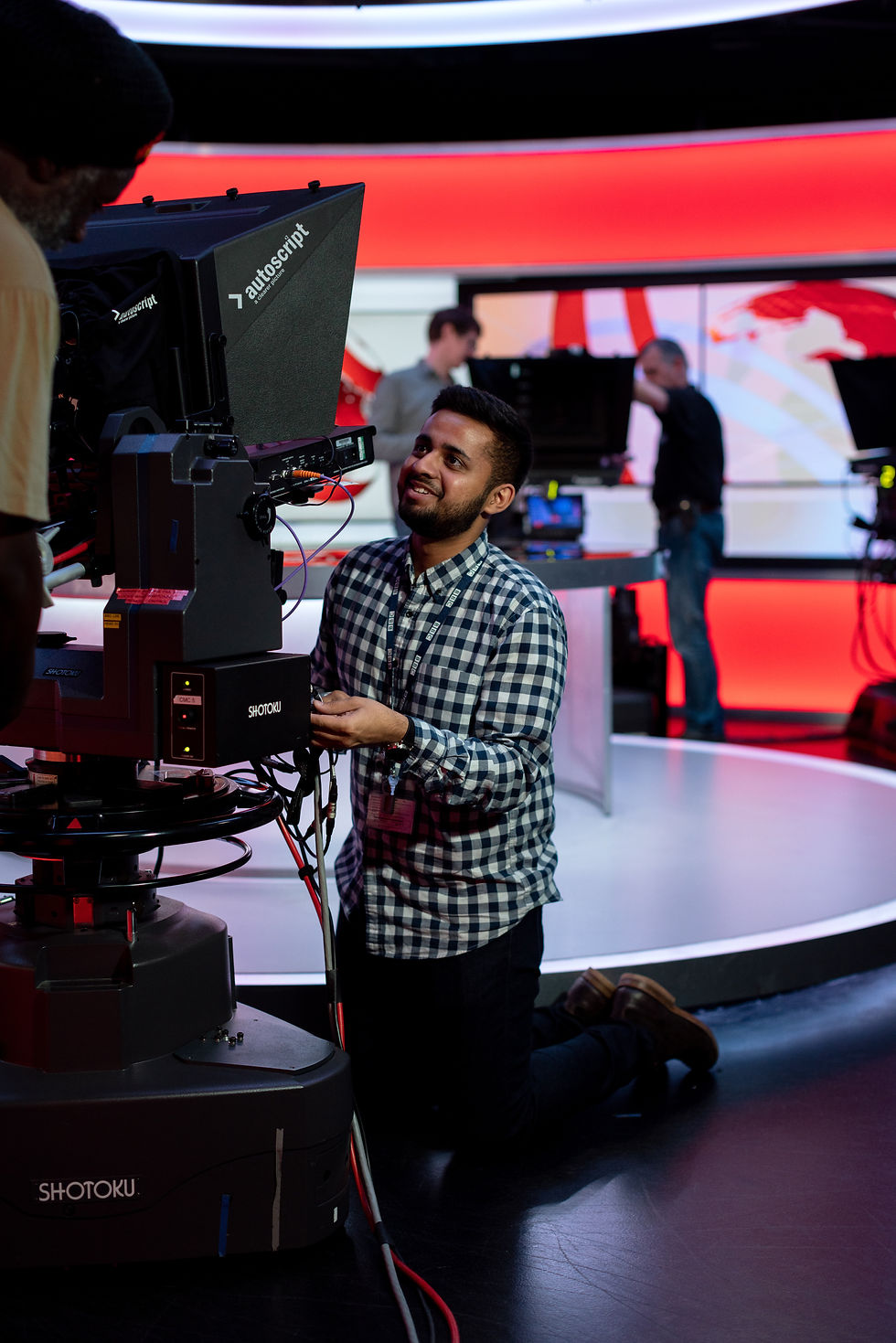 A man in a green and white checked shirt operates some TV broadcast equipment while staff on BBC News work behind him in the studio.