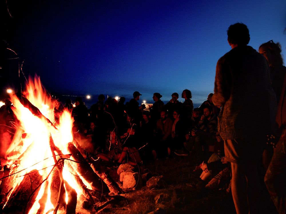 A large bonfire at the beach with a large group of people around