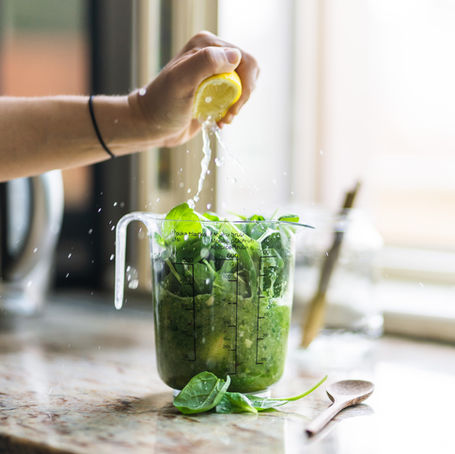 Hand squeezing lemon over a measuring cup filled with spinach on a kitchen counter, water splashing. Sunlight streams through a window.
