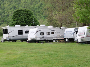 RVs parked on grassy field with a lush forested hill in the background. Trees provide shade, creating a relaxed, nature-filled atmosphere.