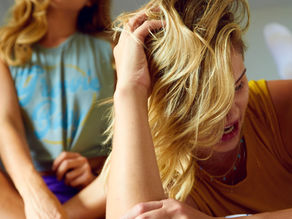 Two women with blond hair on a bed, one in the foreground lying on the bed facing the camera, she is holding her head and looking angry. The other woman is in the background sat on the bed cross legged and also facing the camera out of focus looking upset.