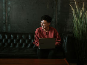 Smiling person with laptop on dark couch, wearing a red sweater in an industrial room with potted grass. Relaxed and cozy vibe.