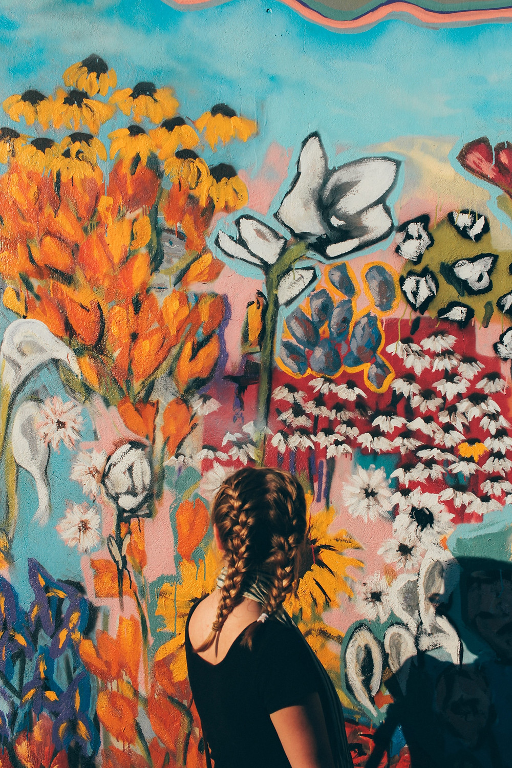 mulher branca com blusa preta olhando para uma parede toda pintada com flores coloridas de todos os tipos possíveis atrás dela.