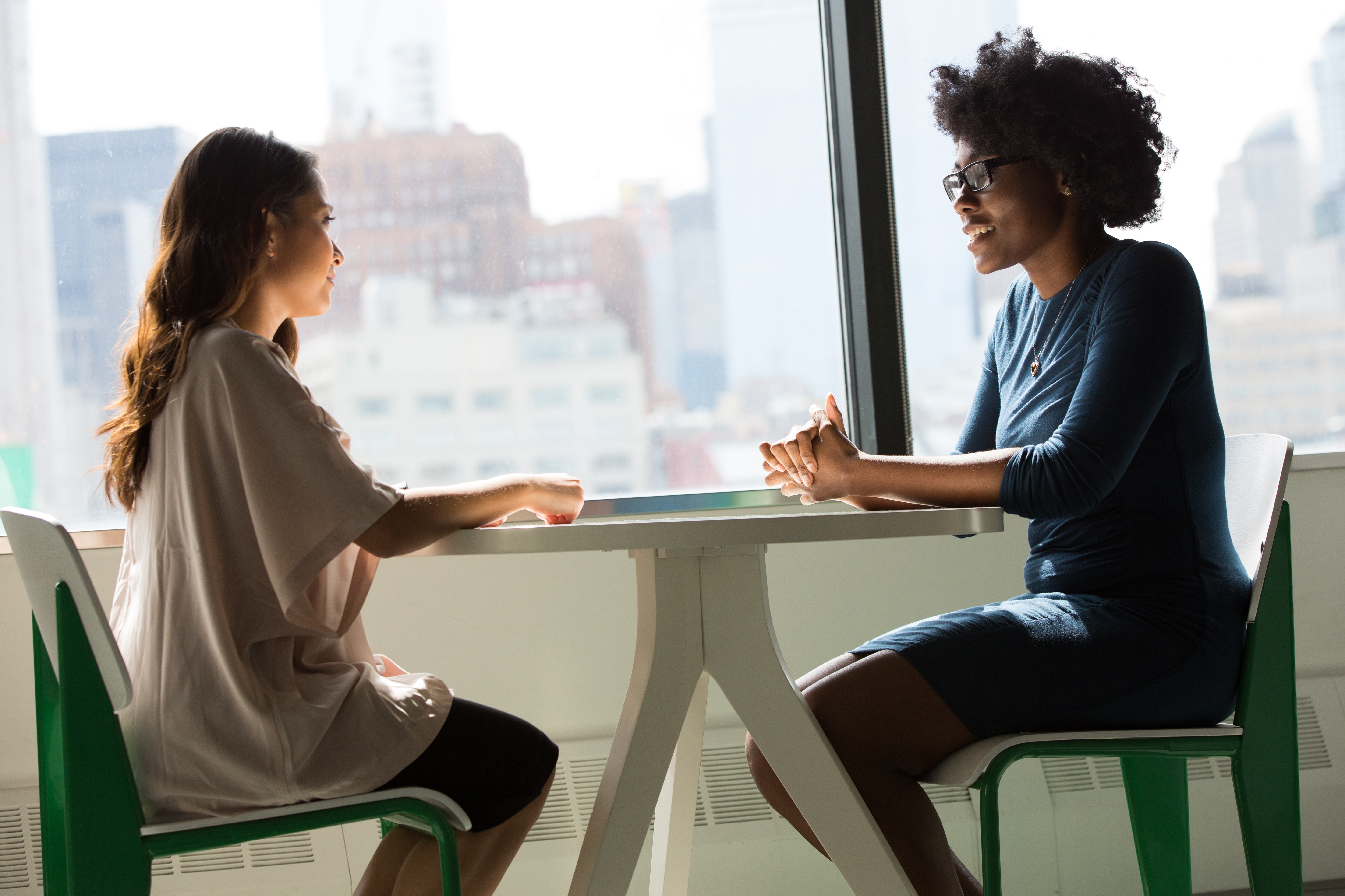 Two business women sit opposite each other a small table next to a window with a blurred cityscape in the background