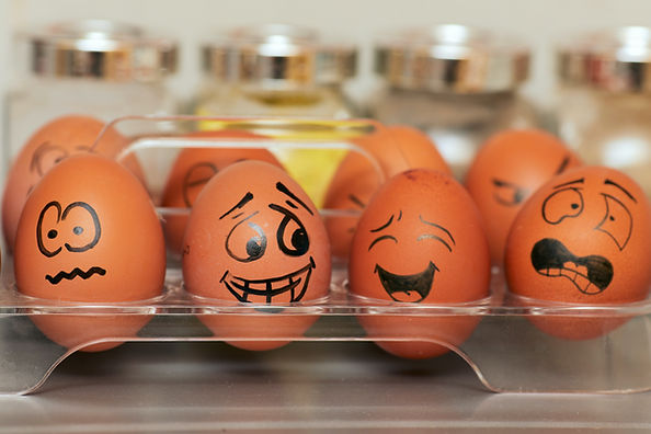 Eggs in a tray with drawn faces showing various emotions, like happy and anxious. Blurred jars in the background. Playful mood.