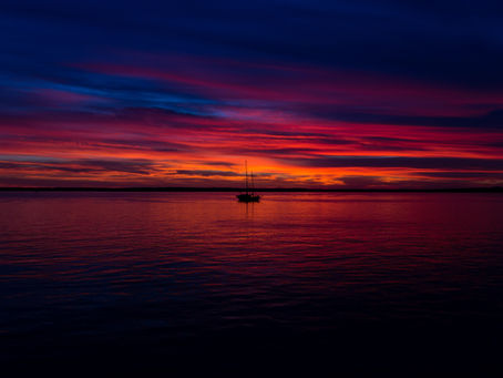 A boat is on the sea as the sun sets, filled with colors of red, pink, blue, and orange in both the sky and the calm sea.