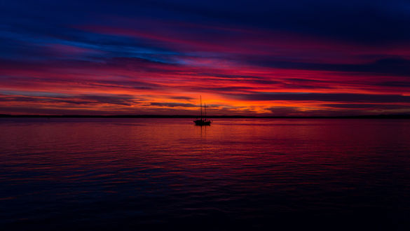 A boat is on the sea as the sun sets, filled with colors of red, pink, blue, and orange in both the sky and the calm sea.