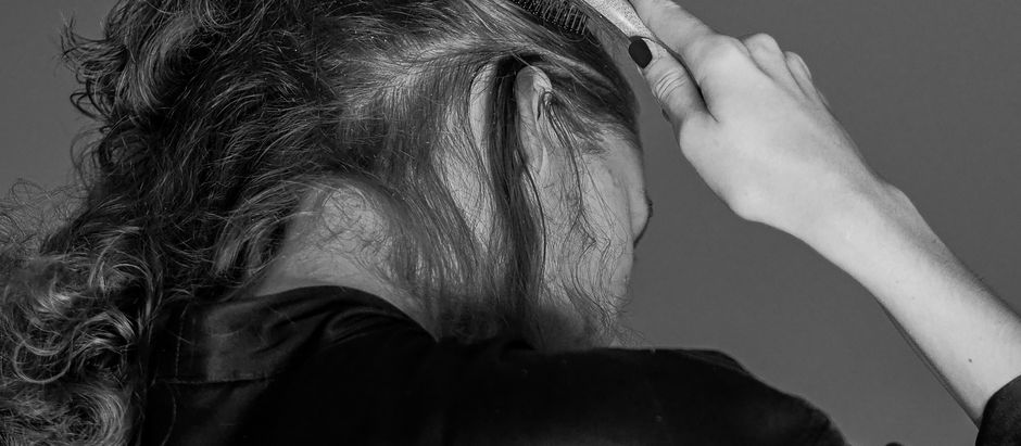 Black-and-white photo of a woman brushing her hair with a comb, seen from behind against a plain background.