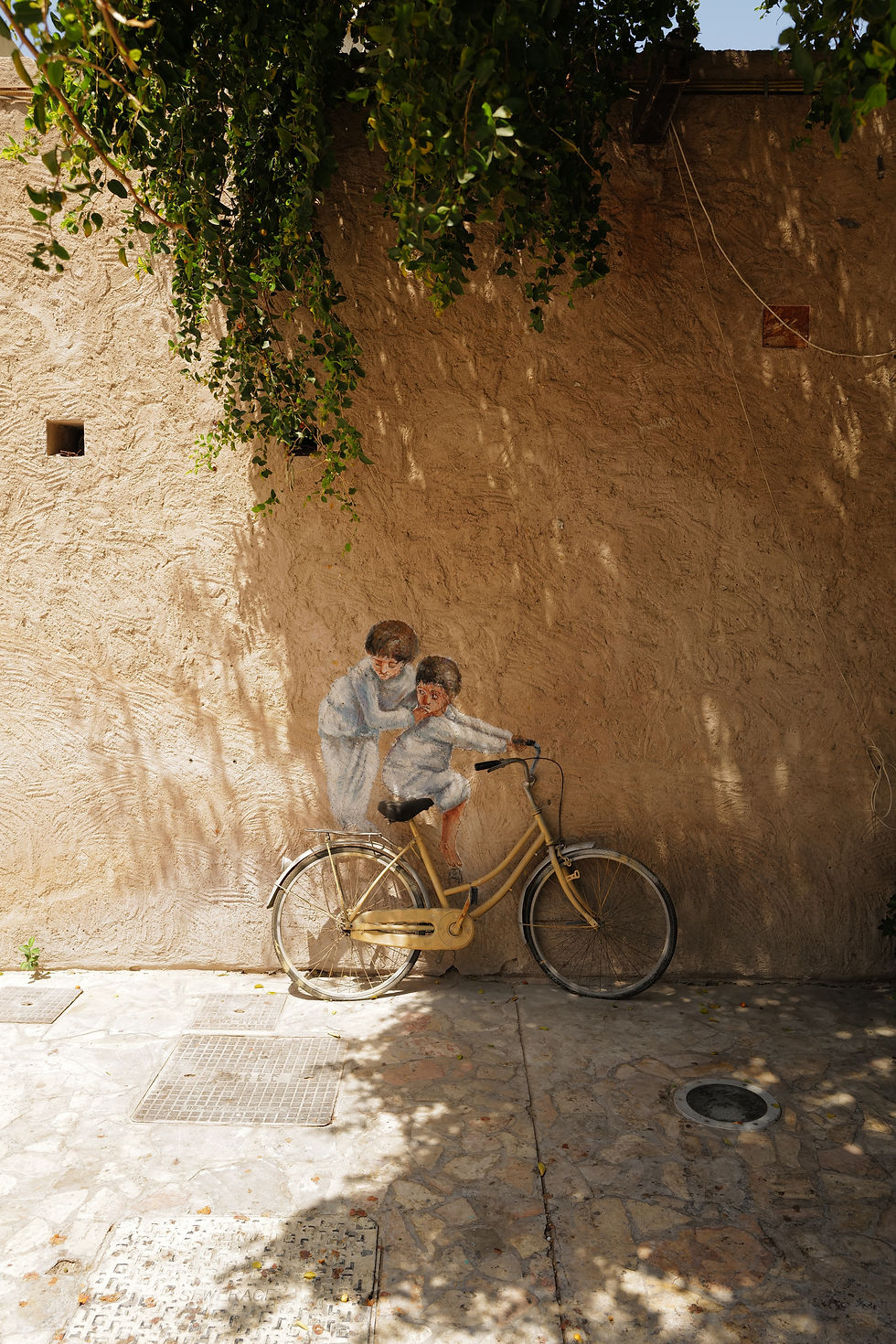 Mural of two children interacting with a bicycle on a textured wall, under green foliage casting dappled shadows on a sunny pavement.