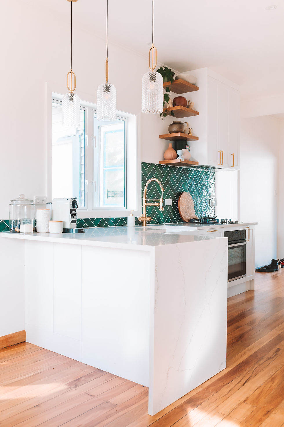 Bright kitchen with white marble counters, green tile backsplash, golden faucet, and wooden shelves holding pottery. Sunlit and airy.