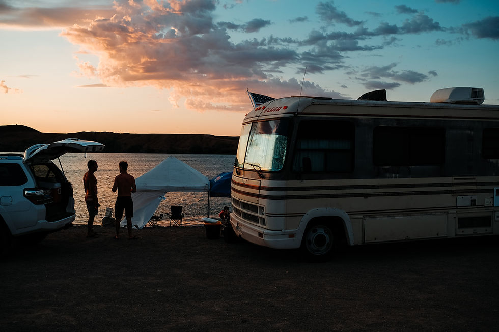 Deux hommes font un camping à côté d'un lac. On voit une voiture sur la gauche et un camping car sur la droite.