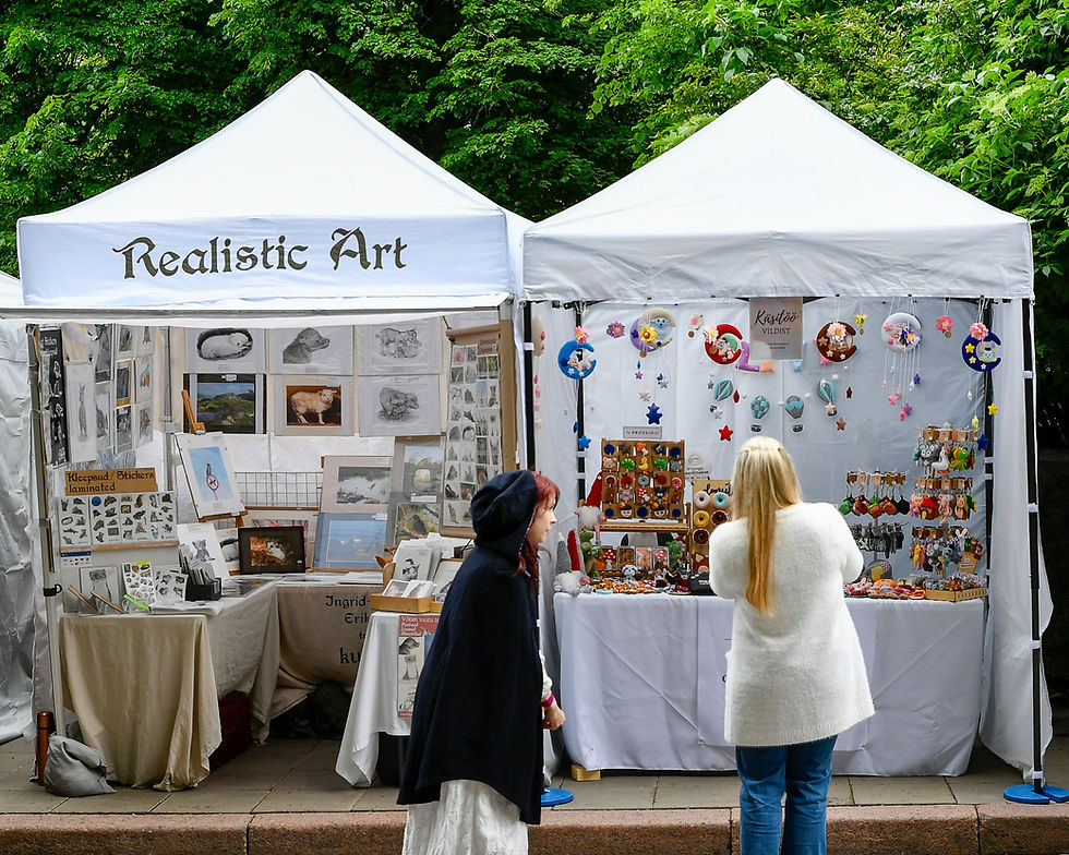 Two women browse art and handmade trinkets at white market tents labeled Realistic Art, under green trees.