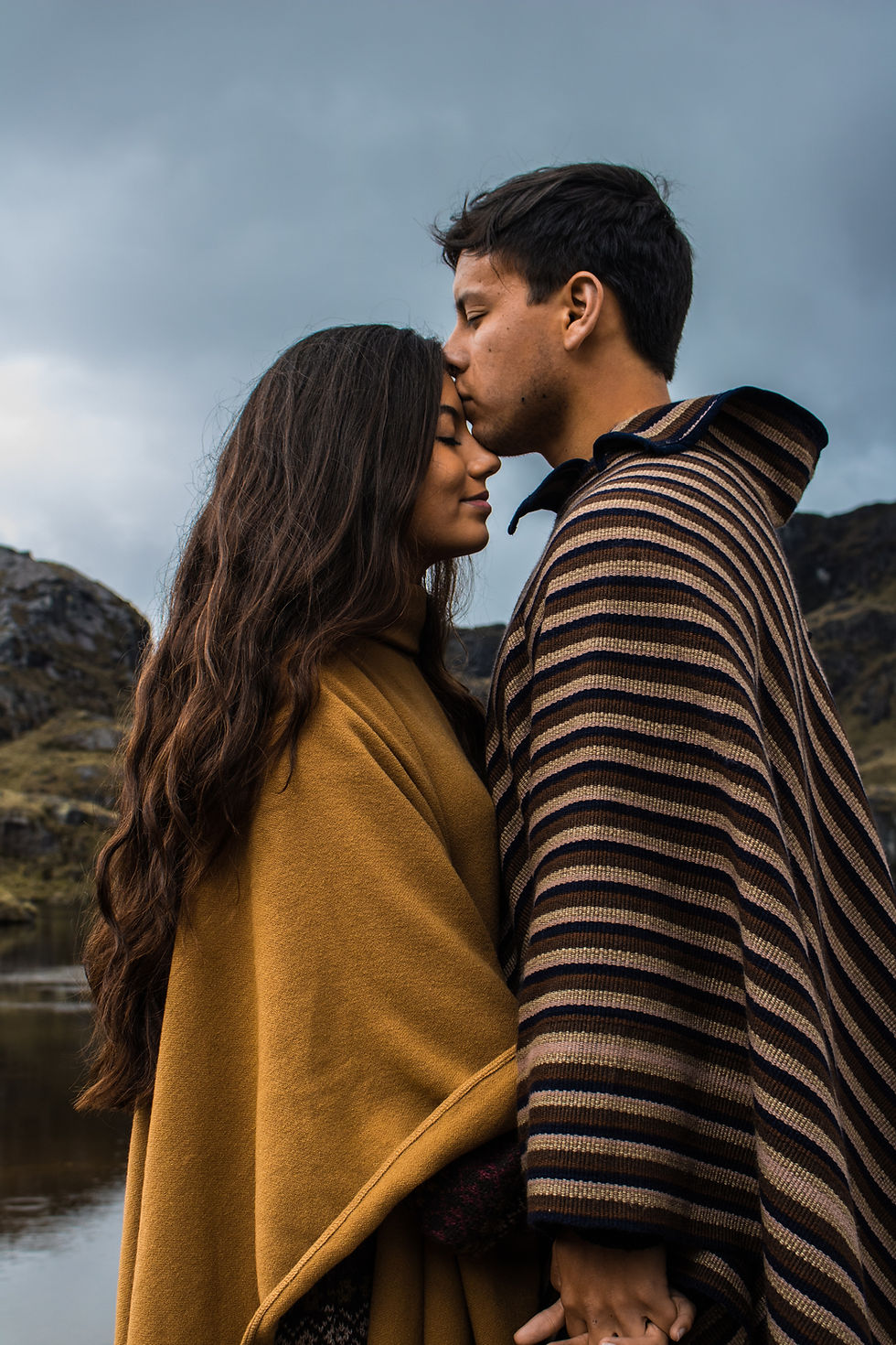 A couple wearing cozy ponchos, embracing in a picturesque South American setting. The man is gently kissing the woman's forehead, capturing a tender moment of love and affection against a backdrop of natural beauty.