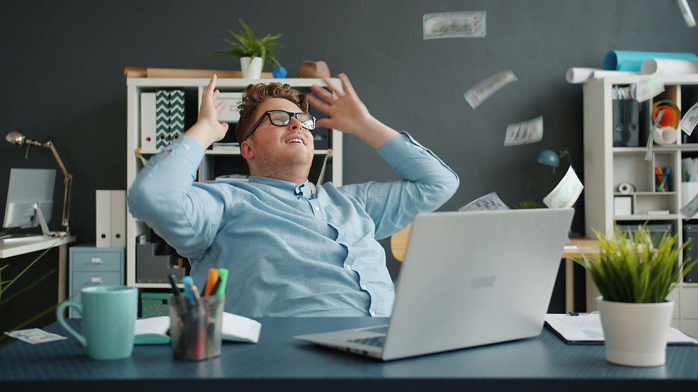 Man in glasses, arms raised, sits at a desk with laptop, papers flying. Shelves with plants and books in the background. Relaxed mood.