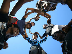 A group of young people stand in a circle, forming a pyramid under a clear blue sky, holding hands and smiling. They convey unity and joy, viewed from below.