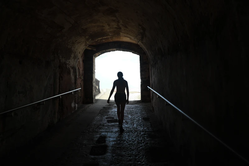 A man standing in a dark room looking out a large window out where there's light