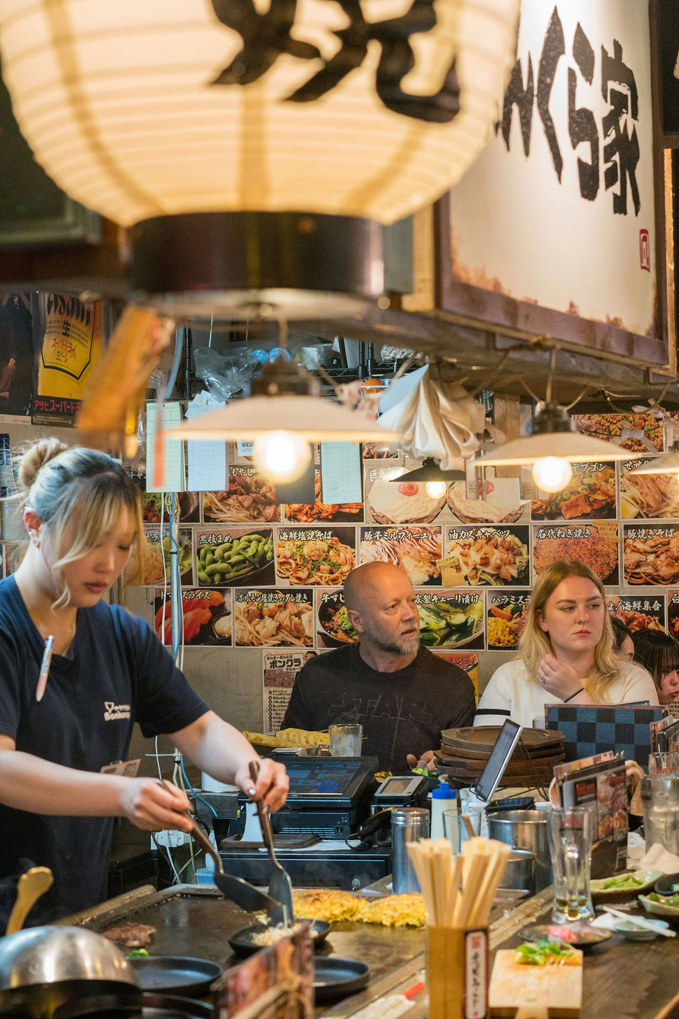 Chef cooking on a grill in a lively, crowded Japanese restaurant. Diners watch attentively. Walls adorned with colorful food images.