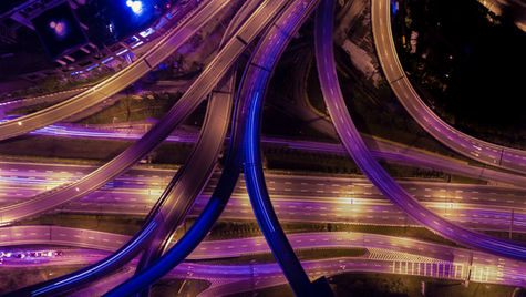 Aerial view of an illuminated highway interchange at night, with curving roads lit in purple and orange, creating a futuristic atmosphere.