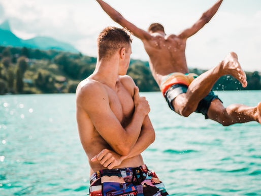 Shirtless man jumps into a turquoise lake while another watches in the foreground, with mountains and bright sky behind.