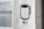 White cloth with a black line drawing of a coffee mug and the word "HELLO" printed beneath. Background has a blurred gray pattern.