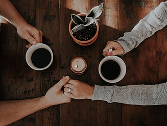 Two hands on a wooden table, one holding a coffee mug, the other clasping it, with a candle and potted plant nearby.