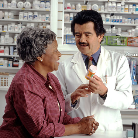 Pharmacist in white coat discusses medication with smiling woman in pink shirt. Shelves of medicine behind them. Friendly and informative mood.